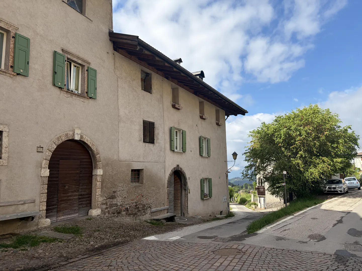 Exterior of a light beige building with green shutters and a large arched wooden door. Cobblestone street and blue sky.