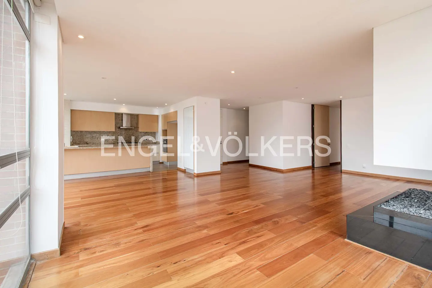 Open-concept living space with hardwood floors, white walls, and a modern kitchen with light wood cabinets. A black fireplace is visible.