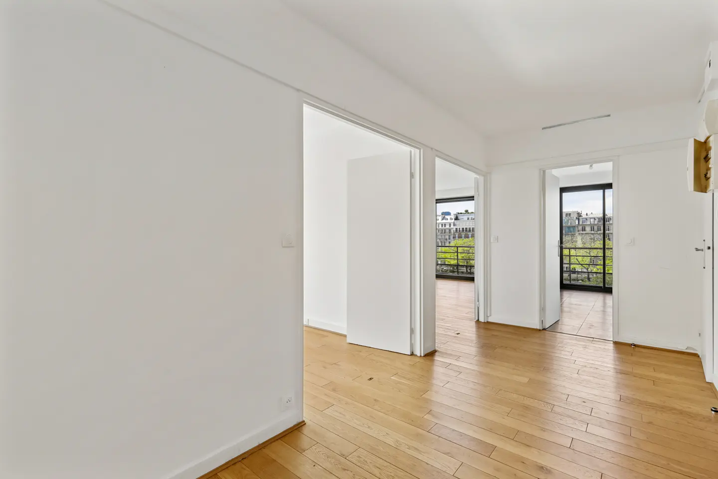 Bright, empty room with white walls and wood floors. Open doorways lead to balconies with city views.
