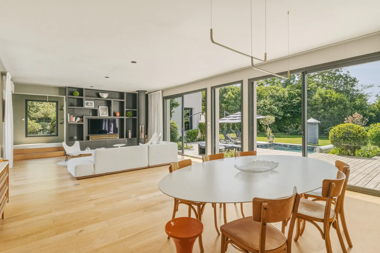Bright, open living space with a white sofa, built-in shelves, and a dining table with wooden chairs. Large windows overlook a pool and lush greenery.