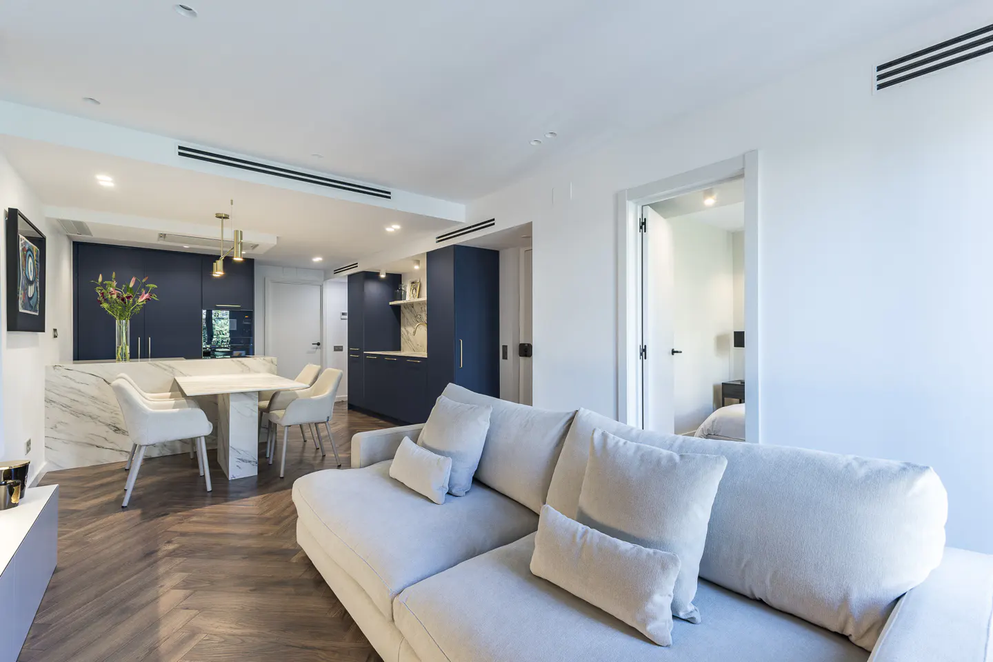 Bright, modern apartment interior with a white sofa, marble dining table, and navy blue kitchen cabinets. Hardwood floors add warmth.