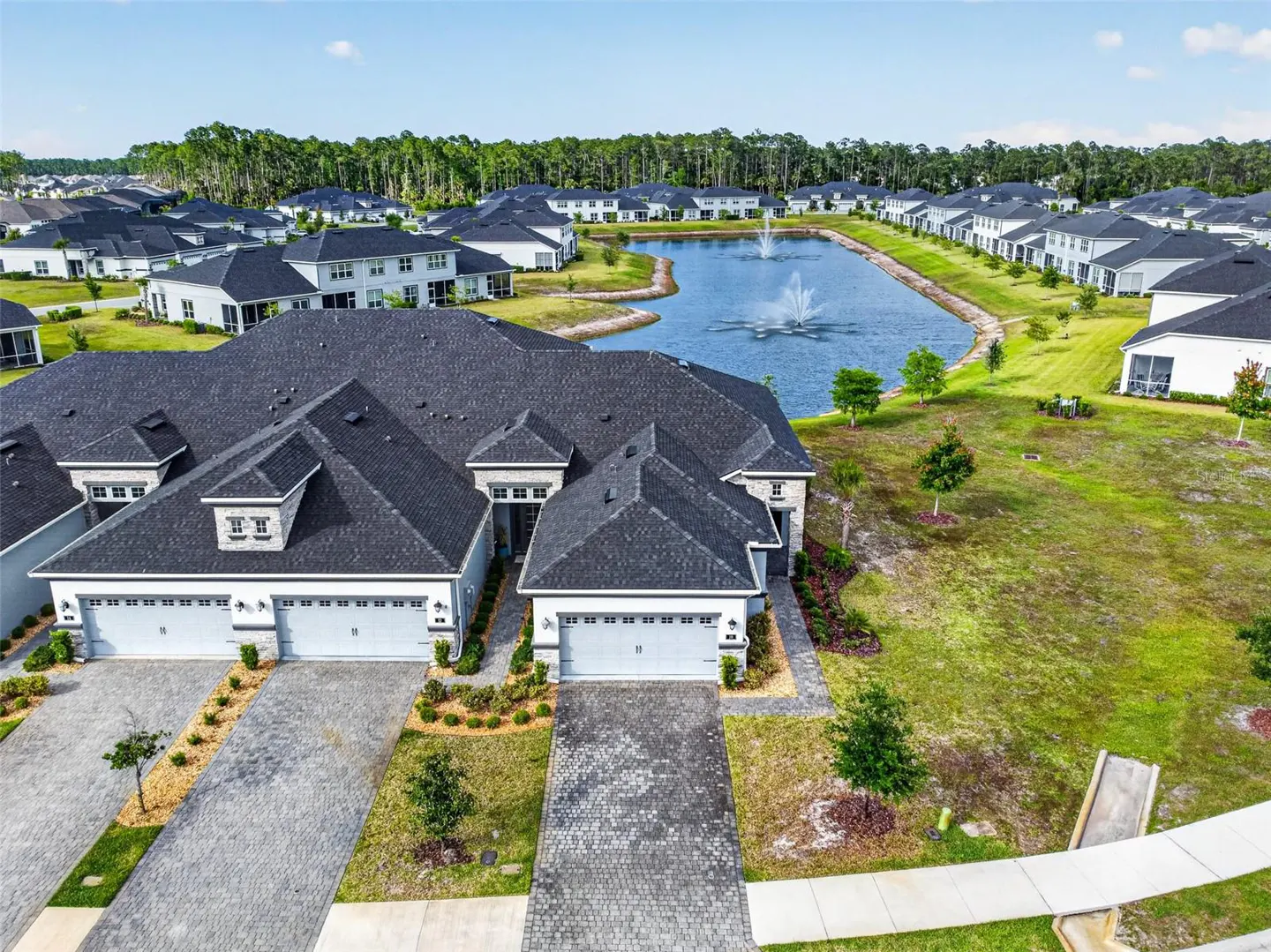 Aerial view of a modern, light-gray home with a dark roof, a pond with a fountain, and other houses in the background.