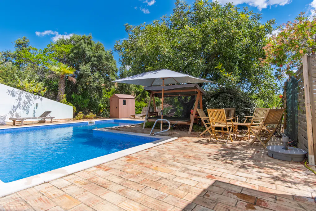 Outdoor pool area with blue water, brick patio, lounge chairs, swing bench under umbrella, and wooden table with chairs.
