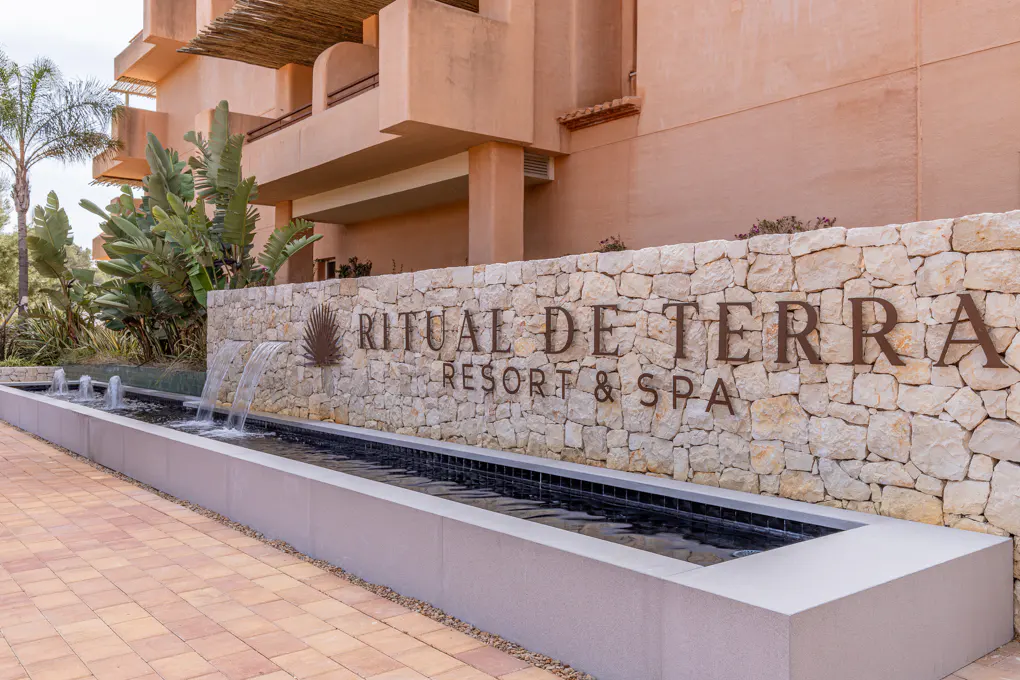 Exterior view of Ritual De Terra Resort & Spa with a stone wall sign and a water feature. The building is a light brown color.