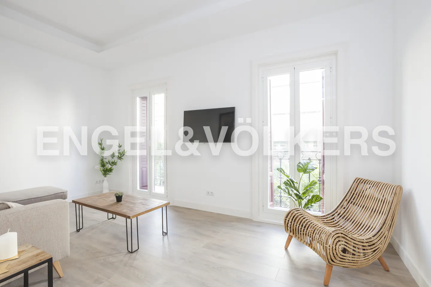 Bright living room with white walls, light wood floors, a rattan chair, coffee table, and a wall-mounted television.