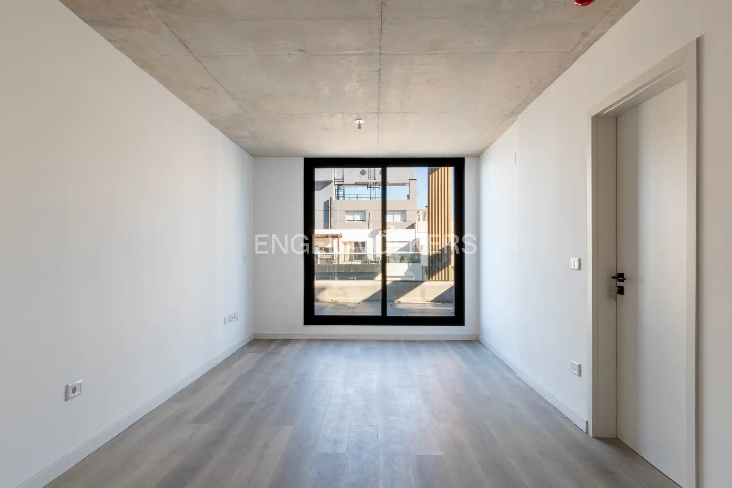 Empty room with white walls, gray wood floor, and concrete ceiling. A black-framed window shows a building exterior. A white door is on the right.
