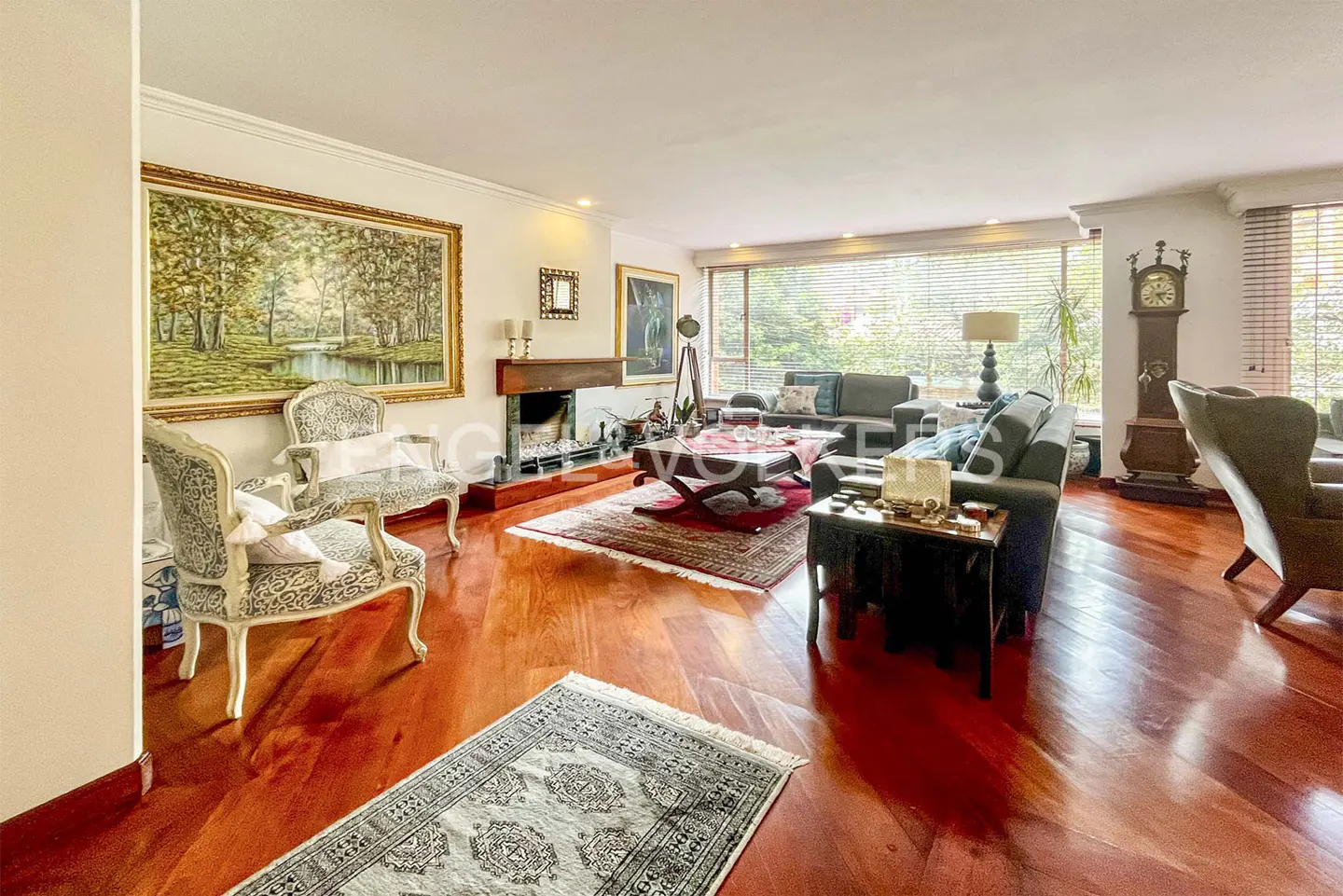 A spacious living room with hardwood floors, a fireplace, sofas, chairs, and a large window with blinds.