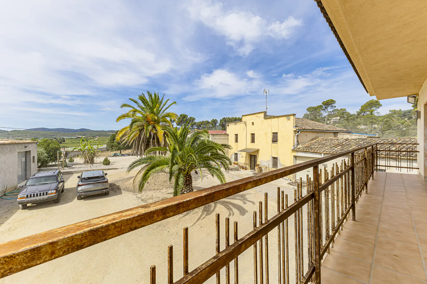 View from a tiled balcony overlooking a sandy courtyard with two parked cars, palm trees, and a yellow rustic building.