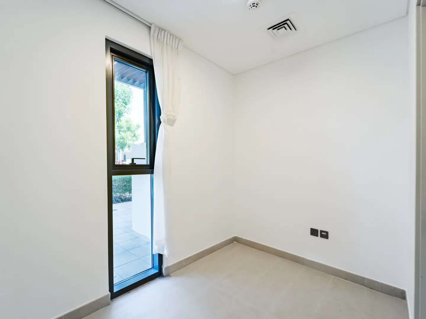 A minimalist room with white walls, tiled floors, and a tall window with a white curtain overlooking a green outdoor area.