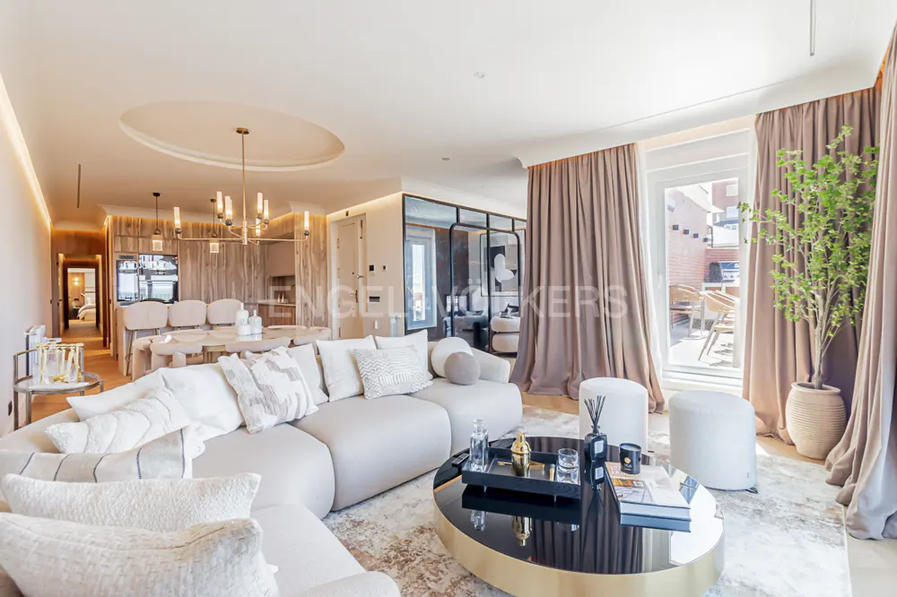 Bright, modern living room with a curved white sofa, black and gold coffee table, and dining area in the background. Curtains frame a balcony view.