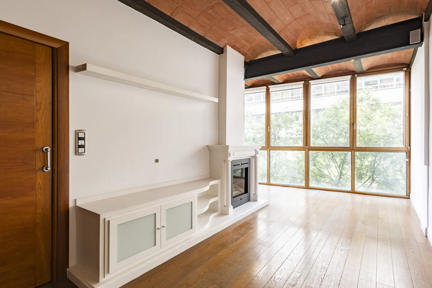 Bright living room with wood floors, white walls, and a fireplace. Large windows offer a view of green trees. A wooden door is on the left.