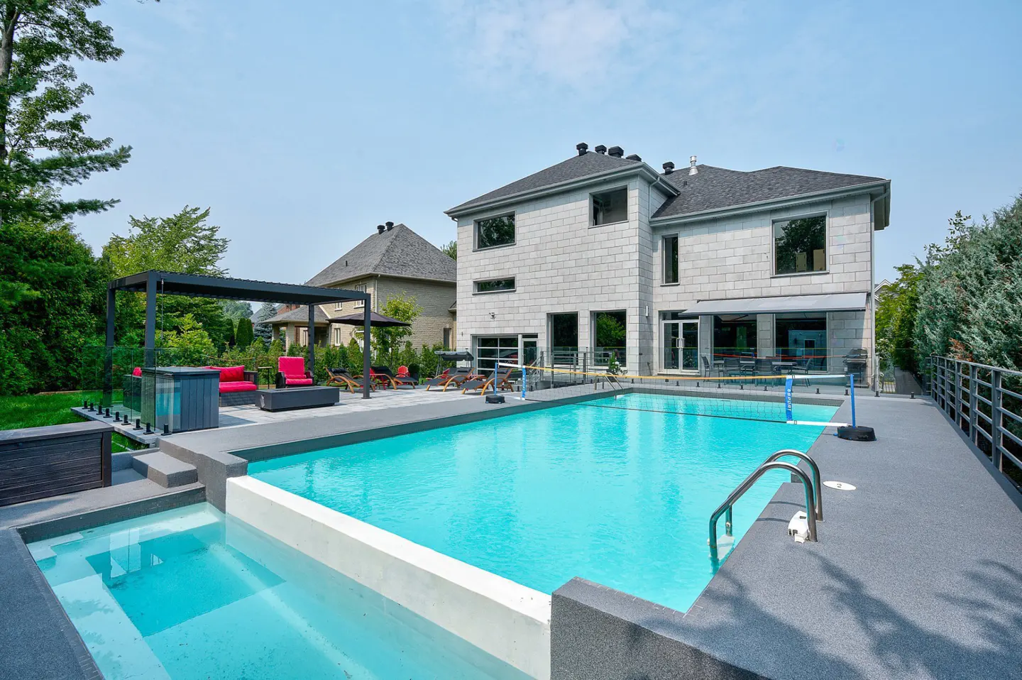 Backyard view of a modern grey stone house with a turquoise pool, lounge area, and green trees.