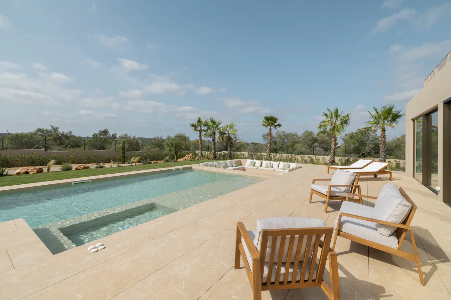 Outdoor patio with pool, lounge chairs, and palm trees under a blue sky. Beige tile flooring and white cushions on the furniture.