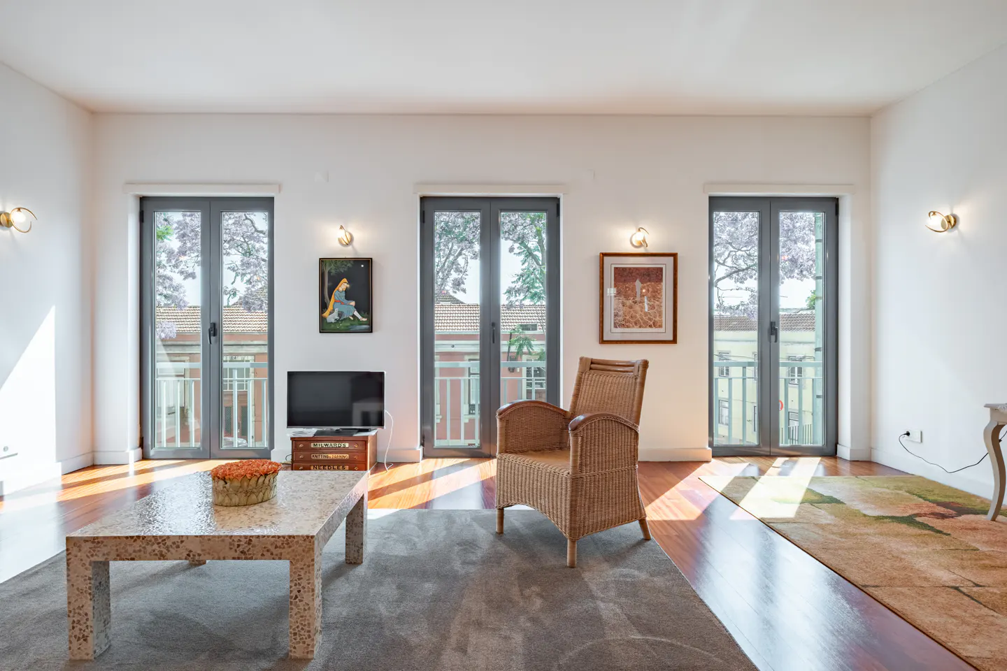 Bright living room with hardwood floors, a wicker chair, a stone coffee table, and three glass doors overlooking a city.