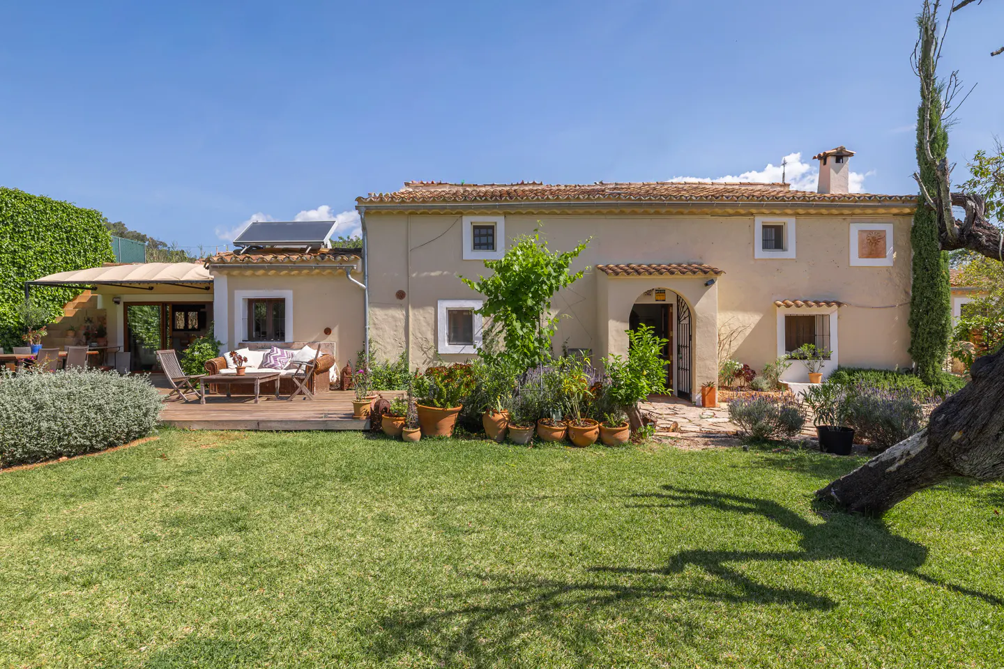 Exterior view of a beige, two-story house with a red tile roof, green lawn, and outdoor seating area on a sunny day.