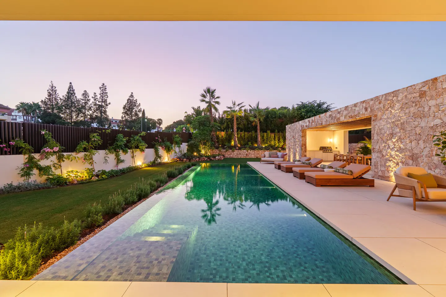 Outdoor pool with lounge chairs and a stone cabana at dusk. Palm trees and green lawn surround the pool.