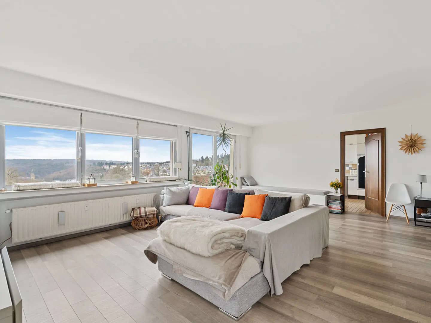 Bright living room with a large window view. A gray sofa with colorful pillows sits on a light wood floor. A doorway leads to a kitchen.