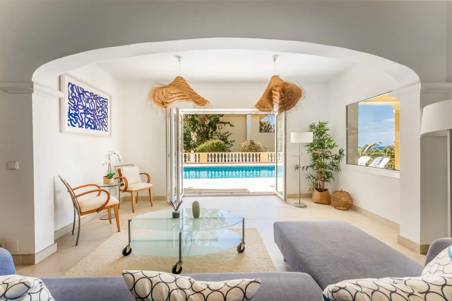 Bright living room with a glass coffee table, grey sofa, and open doors leading to a sunny swimming pool terrace.