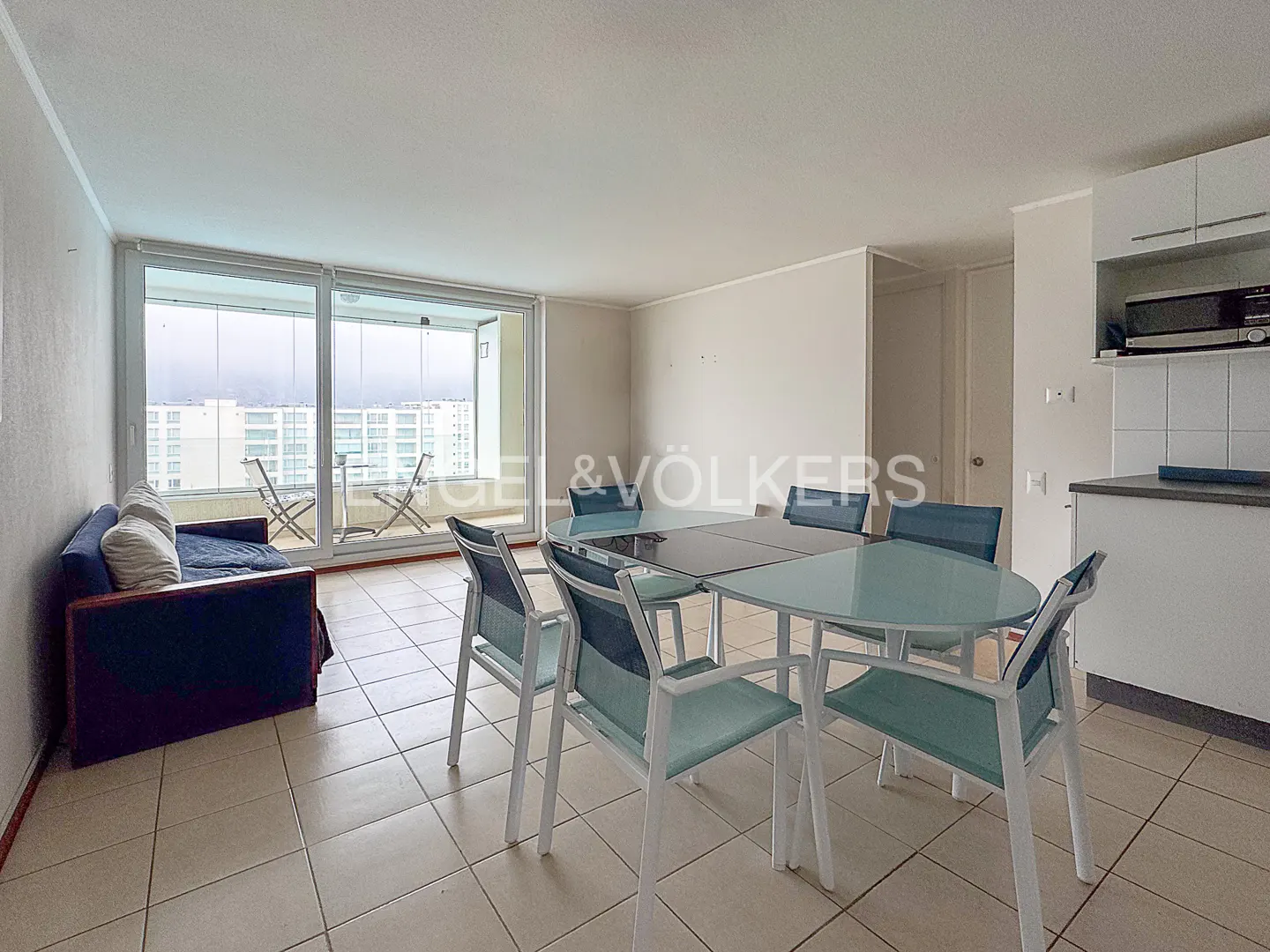 Bright apartment interior with a blue sofa, dining table with six chairs, and a balcony overlooking a cityscape.