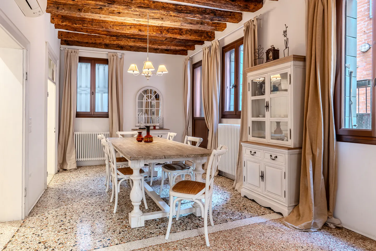 Dining room with rustic wood ceiling beams, terrazzo floors, a wooden table, white chairs, and a white china cabinet.