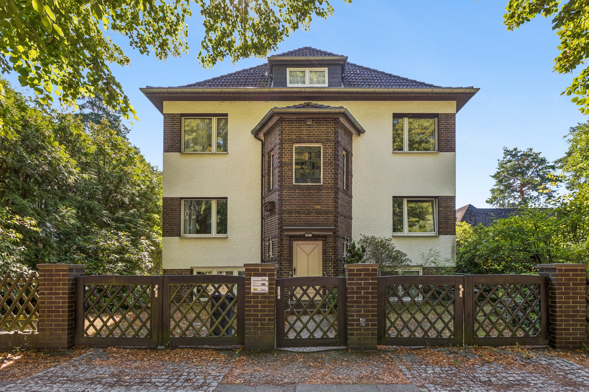 Three-story house with cream stucco and brown brick accents, behind a brown lattice fence, surrounded by lush green trees. [AI-generated text]