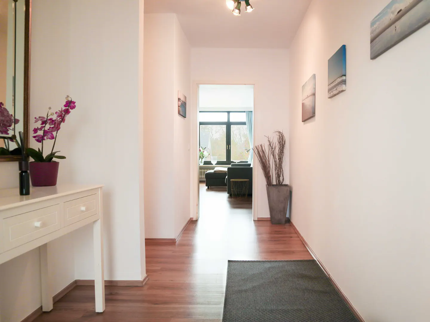 Hallway with wood floors, white walls, and artwork. A white console table with purple orchids sits to the left. A living room is visible in the background.
