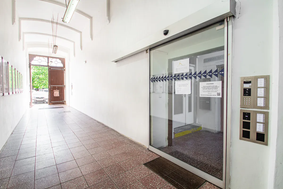 Hallway with tiled floor, white walls, and an open wooden door at the end. A glass sliding door and intercom system are on the right.