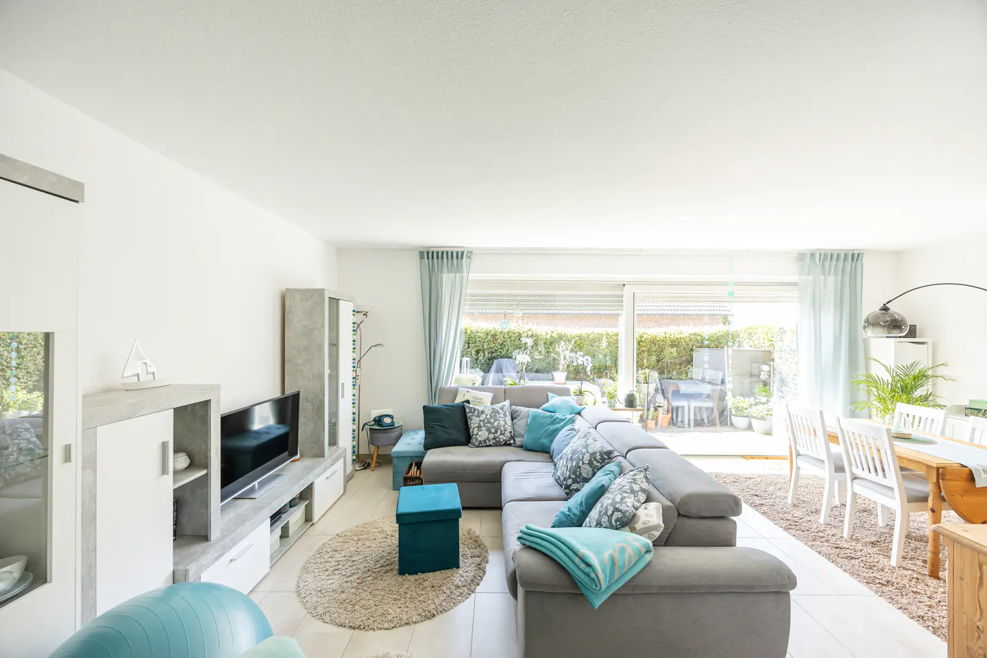 Bright living room with a gray sectional sofa, teal accents, and a dining area near a large window overlooking a green yard.