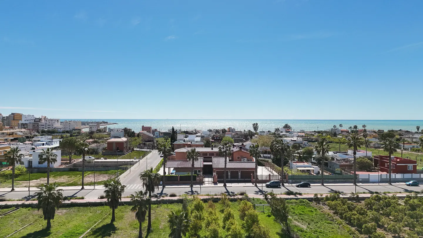 Aerial view of a coastal town with red-roofed houses, palm trees, and the blue sea under a clear sky.
