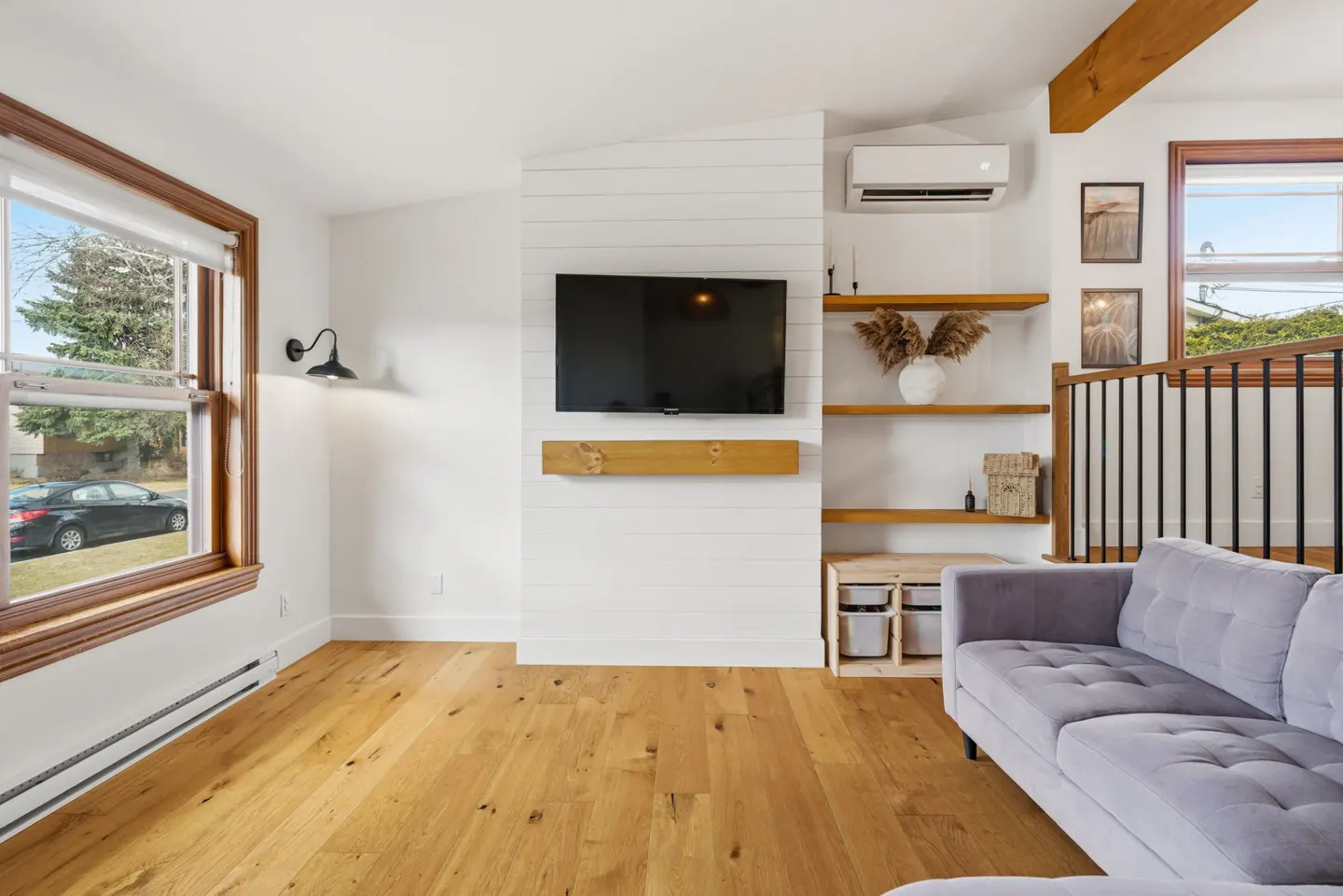 Bright living room with hardwood floors, a gray sofa, and a wall-mounted TV above a wooden shelf. A window shows a car outside.