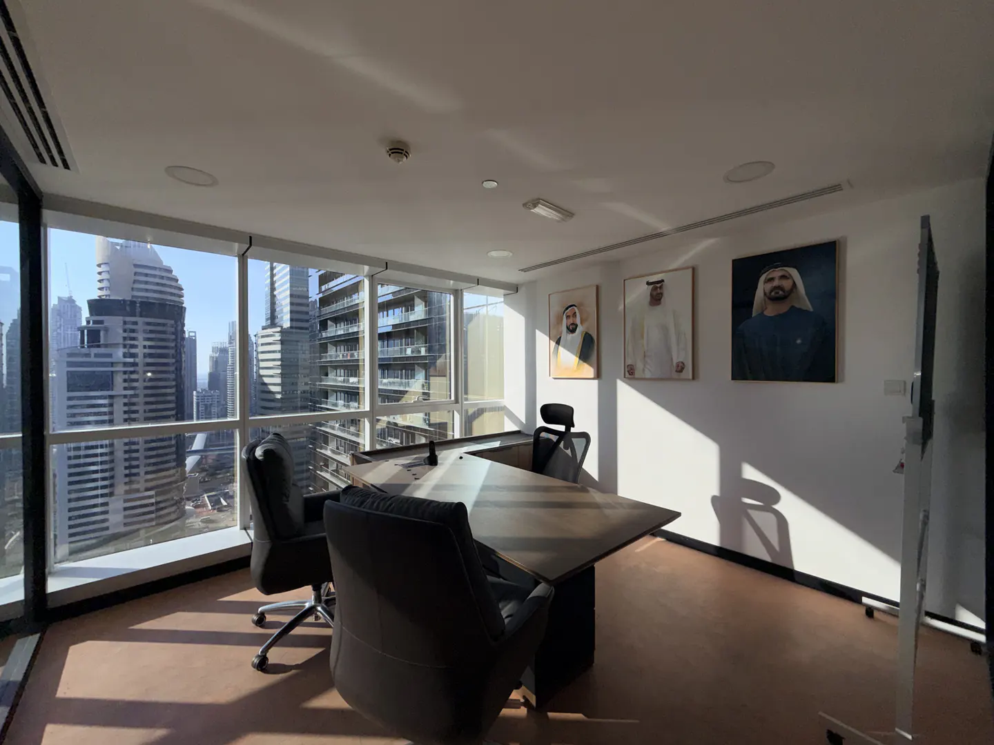 Office space with a large window overlooking a city skyline. Portraits hang on the wall above a dark conference table and chairs.
