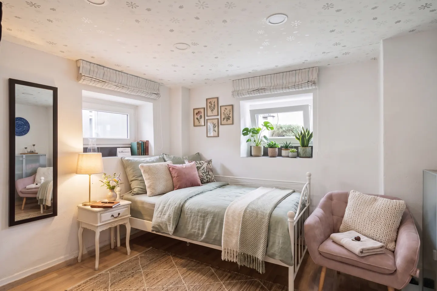 Bedroom with white walls, a white metal bed frame, and a pink chair. Windows have striped blinds and plants. A mirror reflects the room.