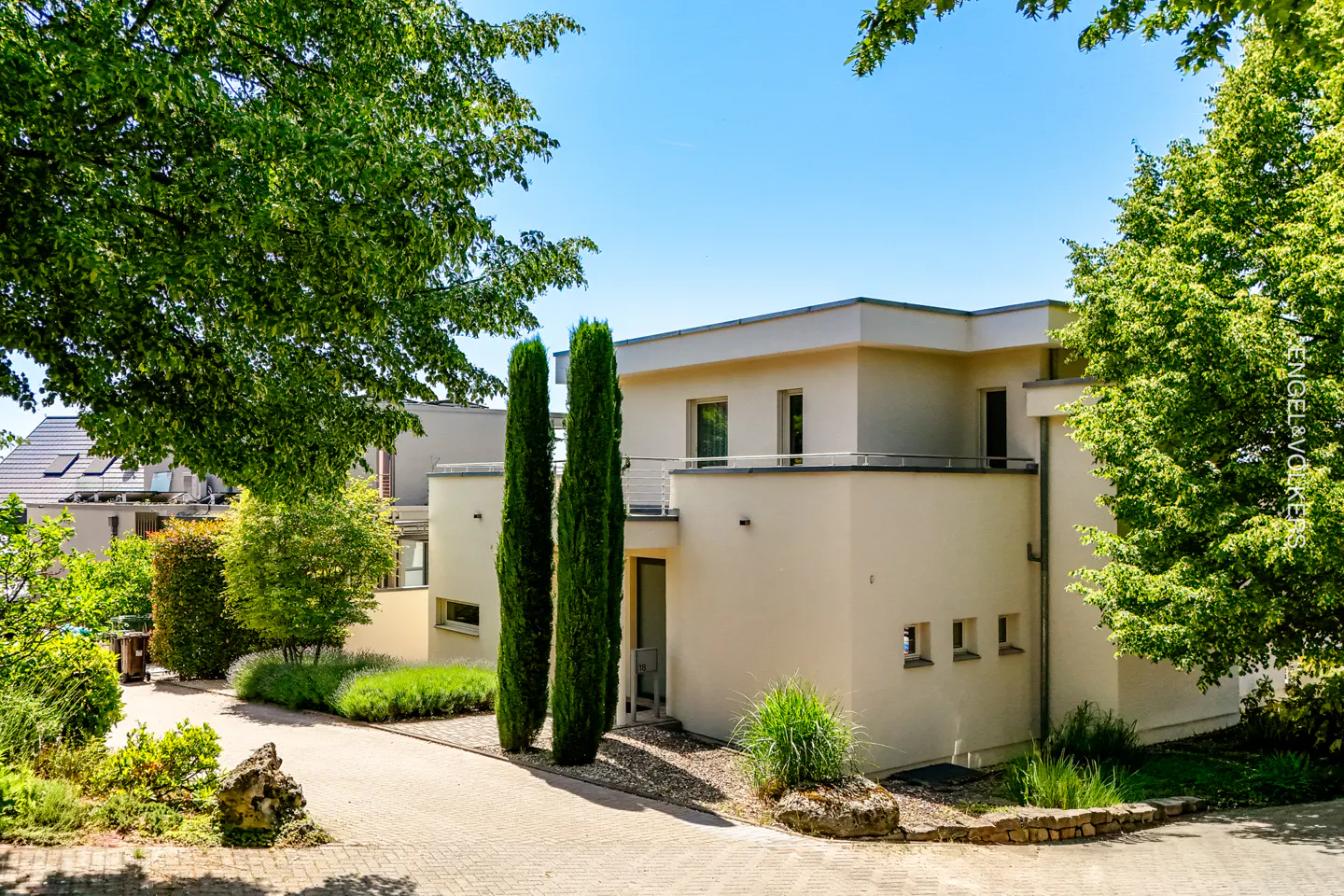 Modern beige two-story house with a paved driveway, manicured landscaping, and tall cypress trees under a clear blue sky.