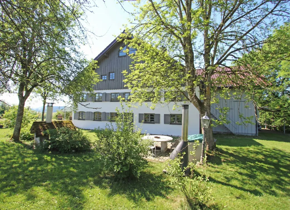 Exterior view of a white two-story house with gray shutters and a gray wood-paneled attic, surrounded by green trees and grass.
