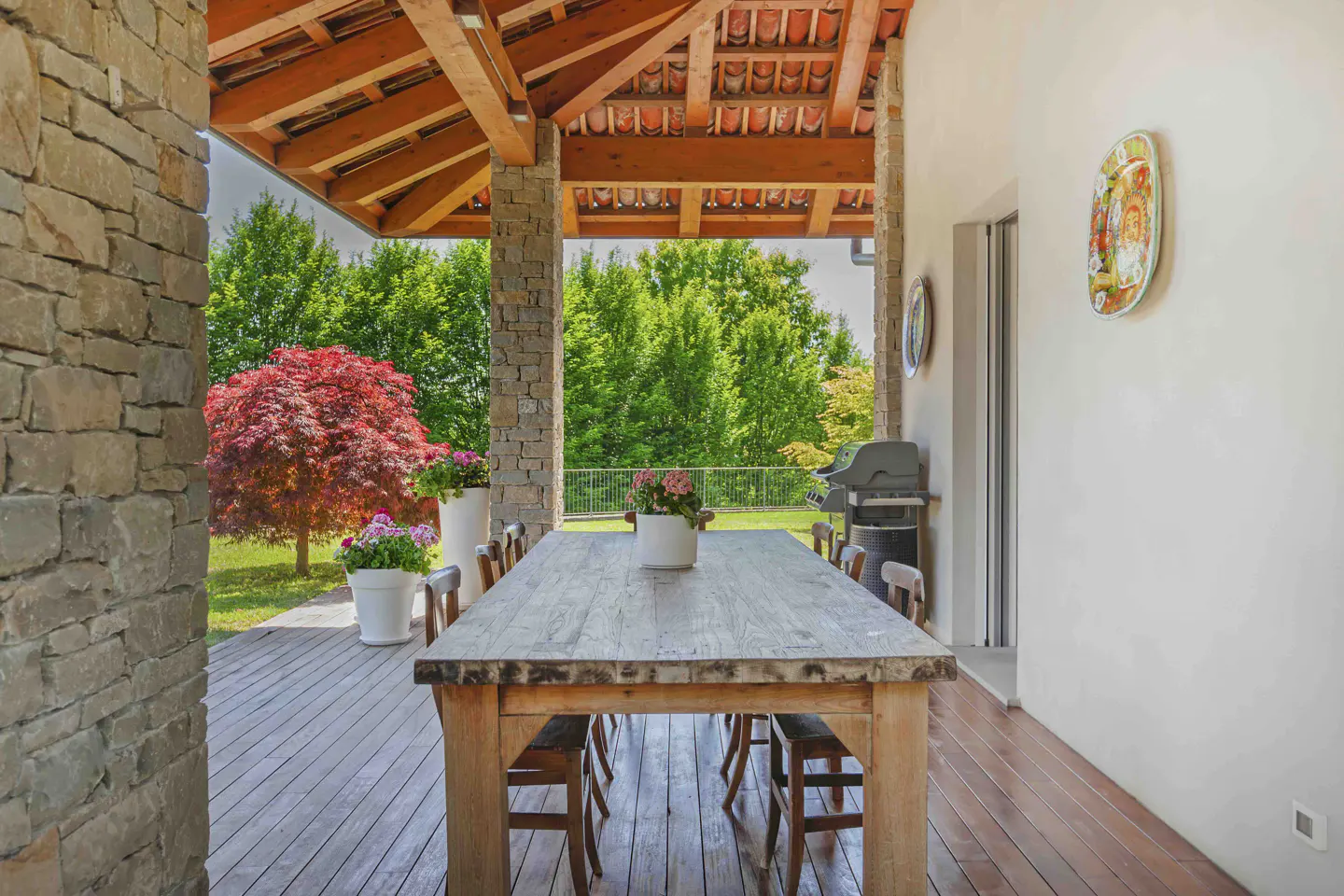 Covered patio with a long wooden table and chairs. Stone columns support a wood beam ceiling. A red maple tree is visible in the yard.