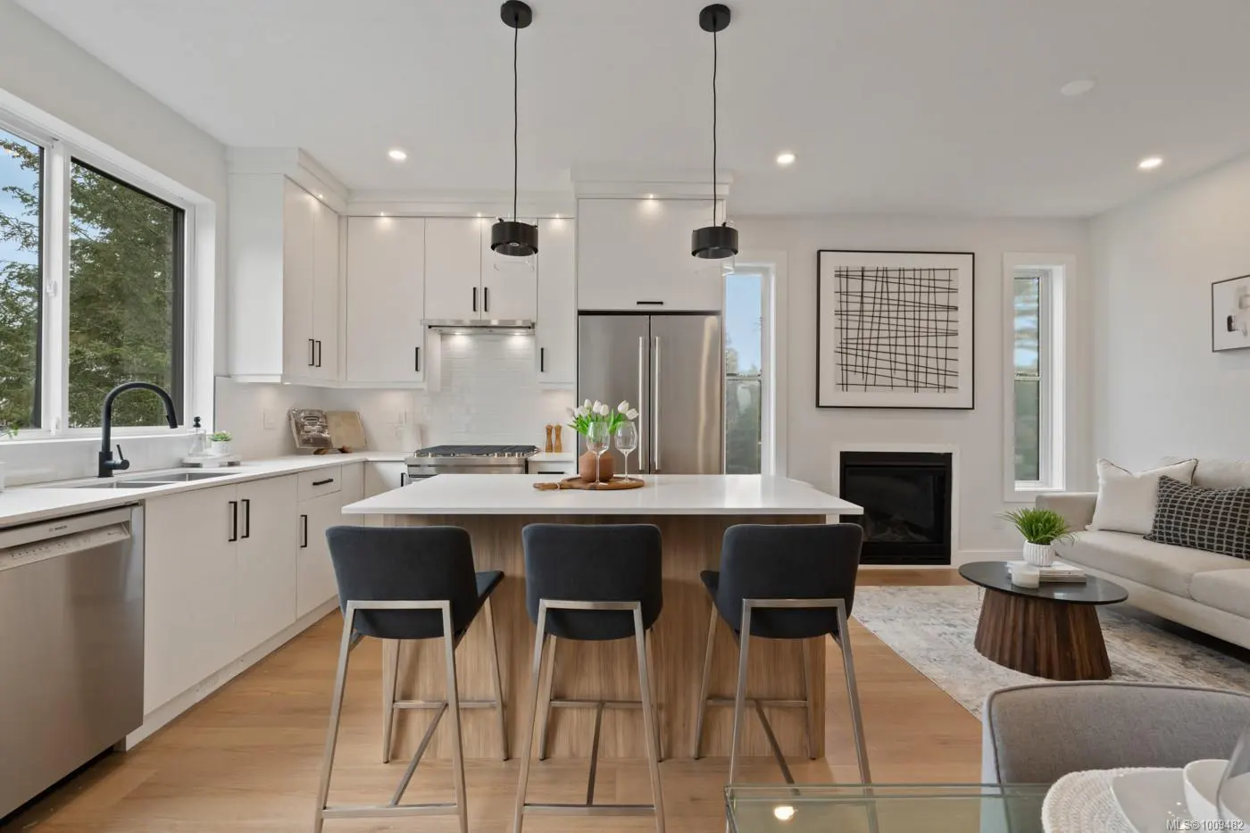 Bright, open-concept kitchen with white cabinets, stainless steel appliances, and a wood-paneled island with three gray bar stools. Living room with fireplace in background.