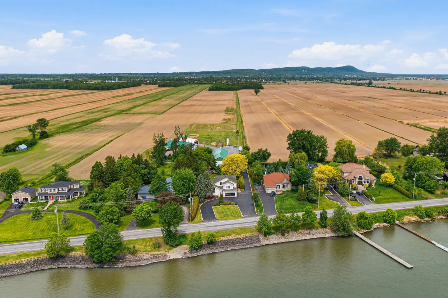 Aerial view of waterfront homes along a road, bordered by vast golden farm fields and distant hills under a blue sky.