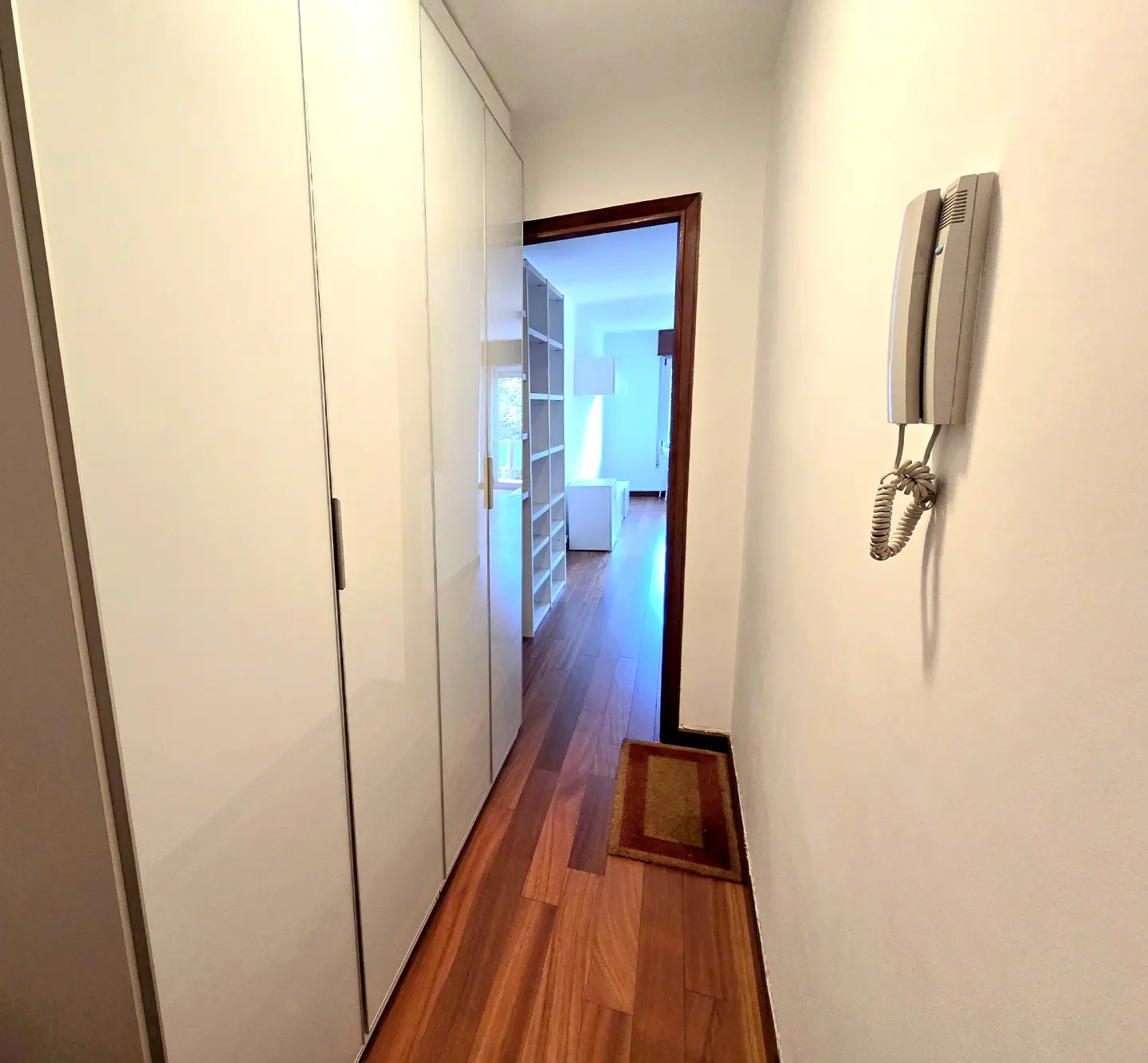 Hallway with wood floors, white walls, and a row of white closets. A doorway leads to a room with white shelving. An intercom is mounted on the wall.