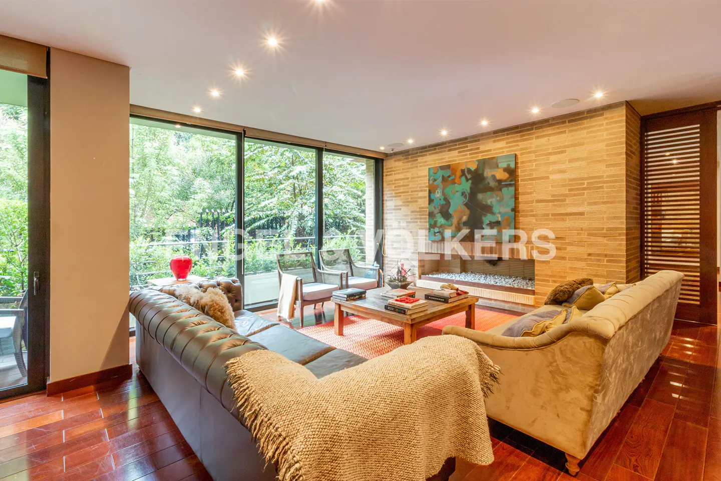 Living room with wood floors, brick wall, fireplace, and large windows overlooking greenery. Sofas and chairs provide seating.