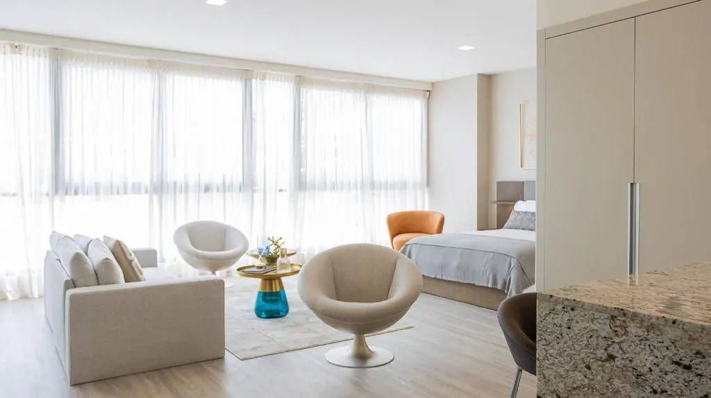 Bright studio apartment with a sofa, chairs, and a bed visible through an opening. Large windows with sheer white curtains let in natural light.