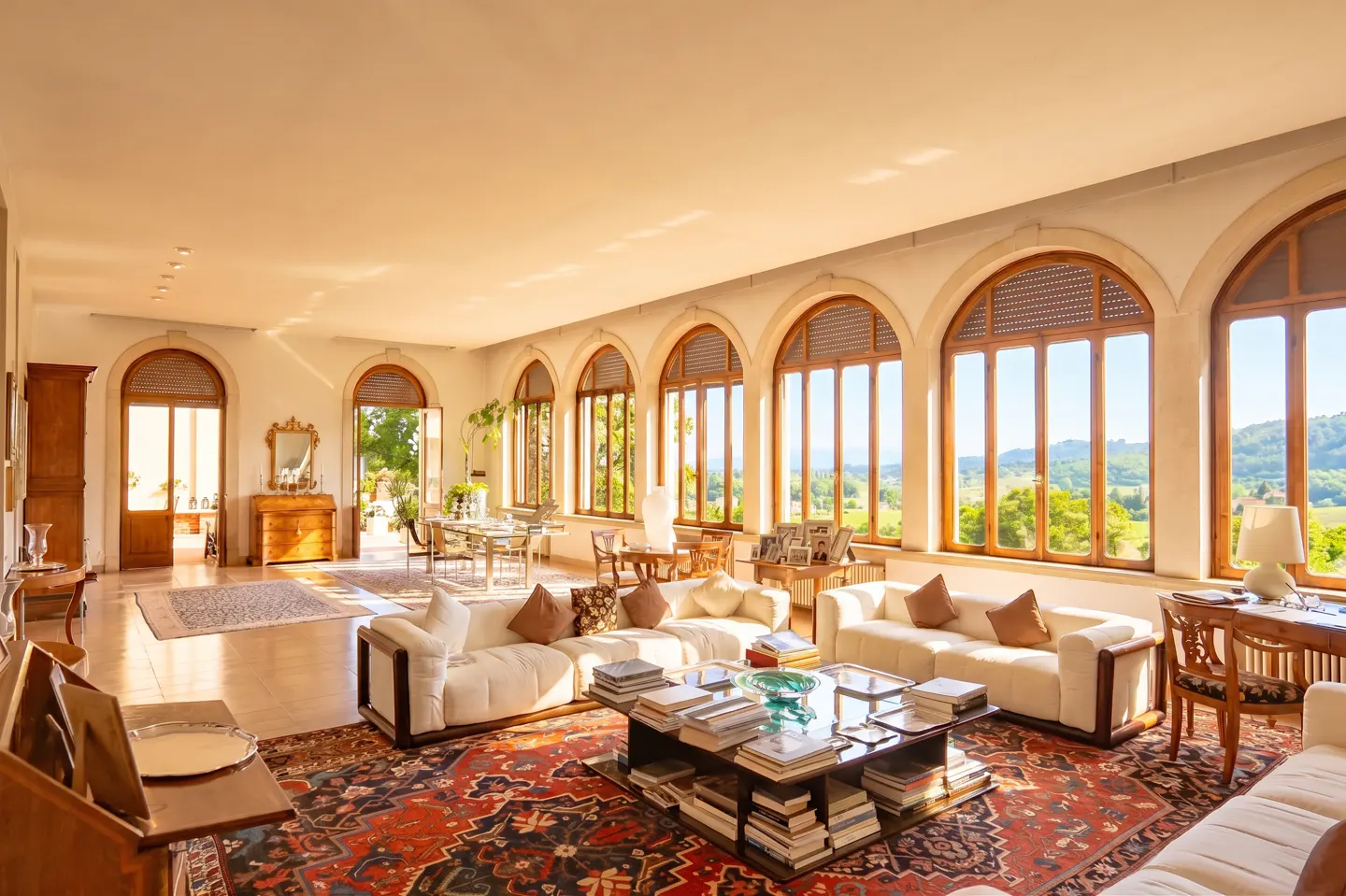 Bright, airy living room with white sofas, a book-filled coffee table, and arched windows overlooking a green landscape.