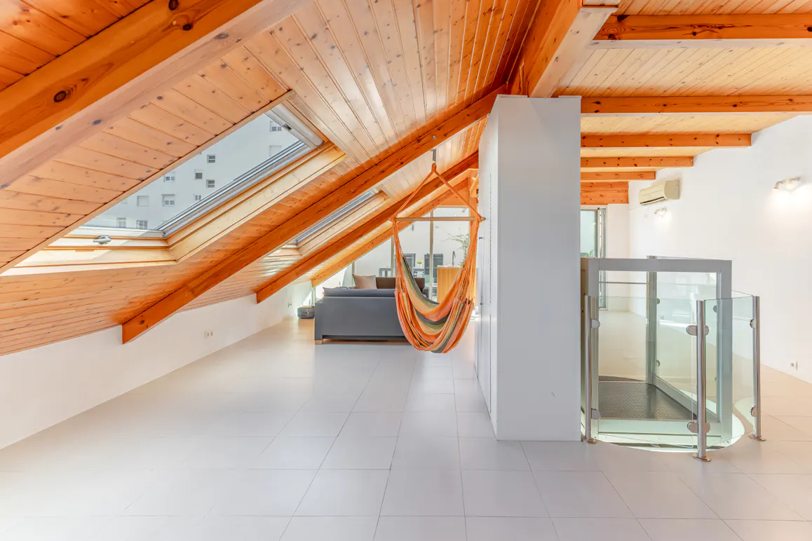 Attic room with wood ceiling, skylight, and white tile floor. A colorful hammock hangs near a glass-enclosed elevator. A gray sofa is in the background.