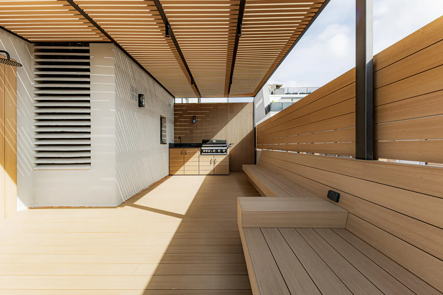 Outdoor patio with wood benches, grill, and shower. The patio has a wood slat roof and a white wall with vents.