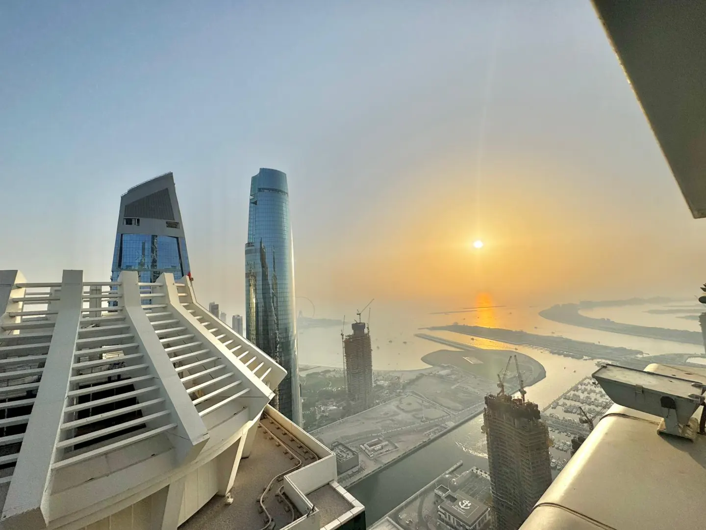 View from a high-rise balcony overlooking a city skyline at sunset. Modern buildings, water, and construction sites are visible.
