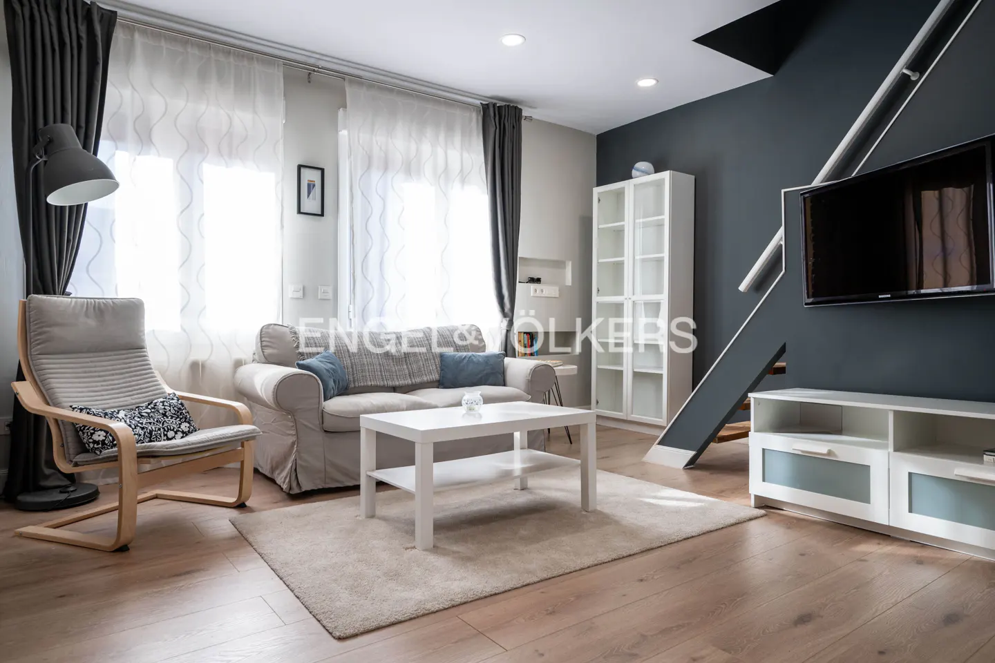 Living room with a beige sofa, white table, and a gray accent wall with a TV and stairs.
