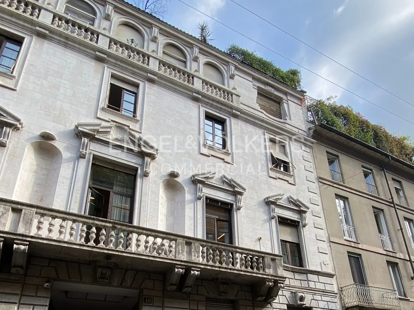 Exterior of a light-colored building with dark-framed windows and a stone balcony. Greenery is visible on the roof.