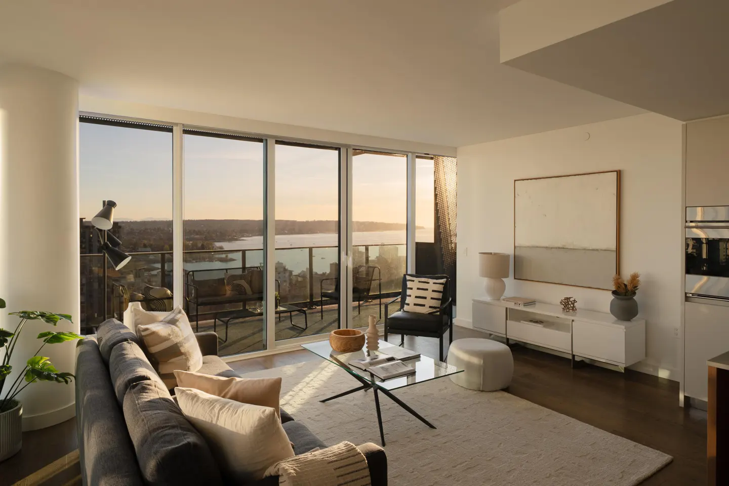 Bright, modern living room with a gray sofa, glass coffee table, and balcony overlooking a sunset view of water and trees. Artwork and a white console table are visible.
