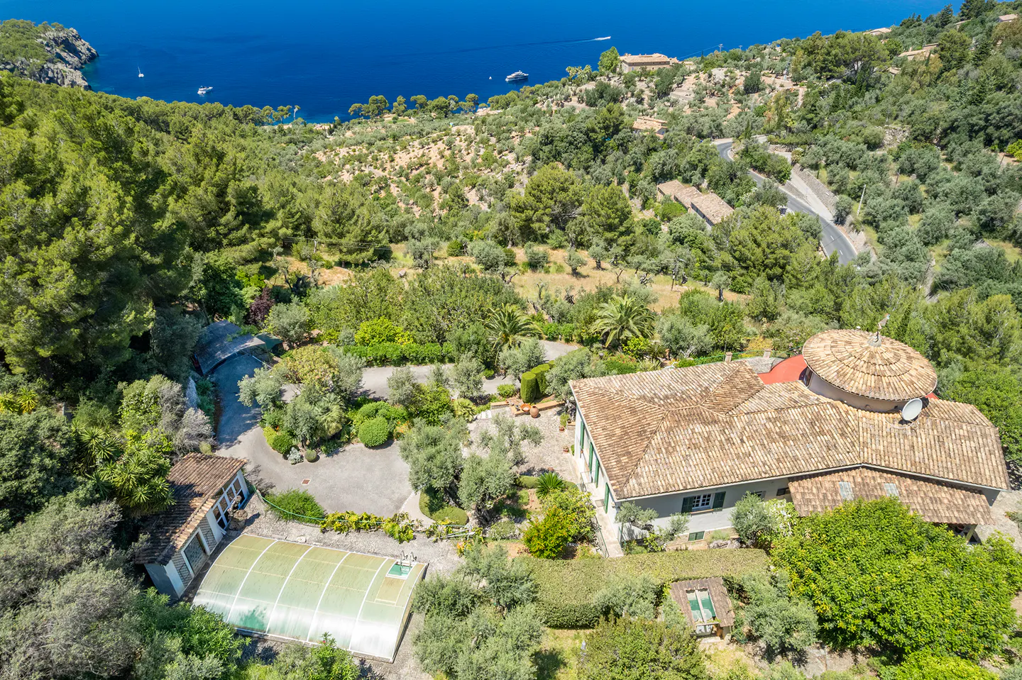 Aerial view of a Mediterranean villa with a tiled roof, pool enclosure, and lush gardens overlooking the blue sea.
