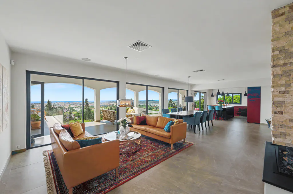 Bright, open-concept living space with tan sofas, blue accents, and a large rug. Sliding glass doors lead to a balcony with a scenic view.