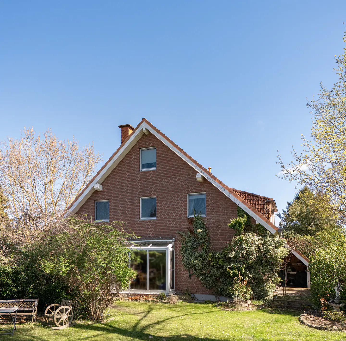A two-story brick house with a red roof and white trim, surrounded by green grass and trees under a blue sky.